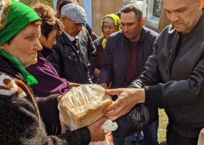 A man hands a loaf of bread to an elderly woman while others wait in line outdoors, conveying a sense of community support.