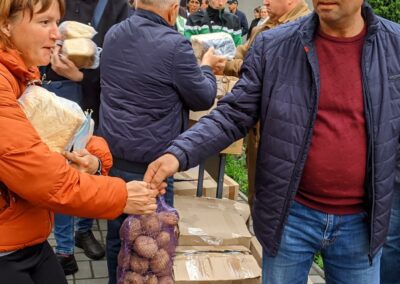 A woman hands a bag of potatoes to a man at an outdoor food distribution event with several people waiting in line.