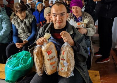 A man sitting on a bench inside a room, holding two bags of bread, surrounded by a crowd of people.