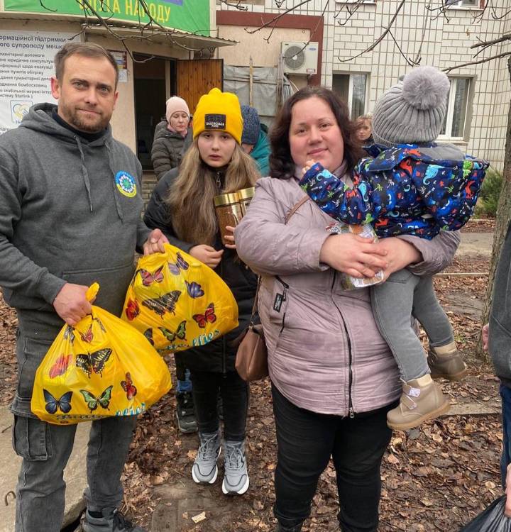 A family stands outside a building, holding bags of supplies and a small child, with a sign in cyrillic text in the background.