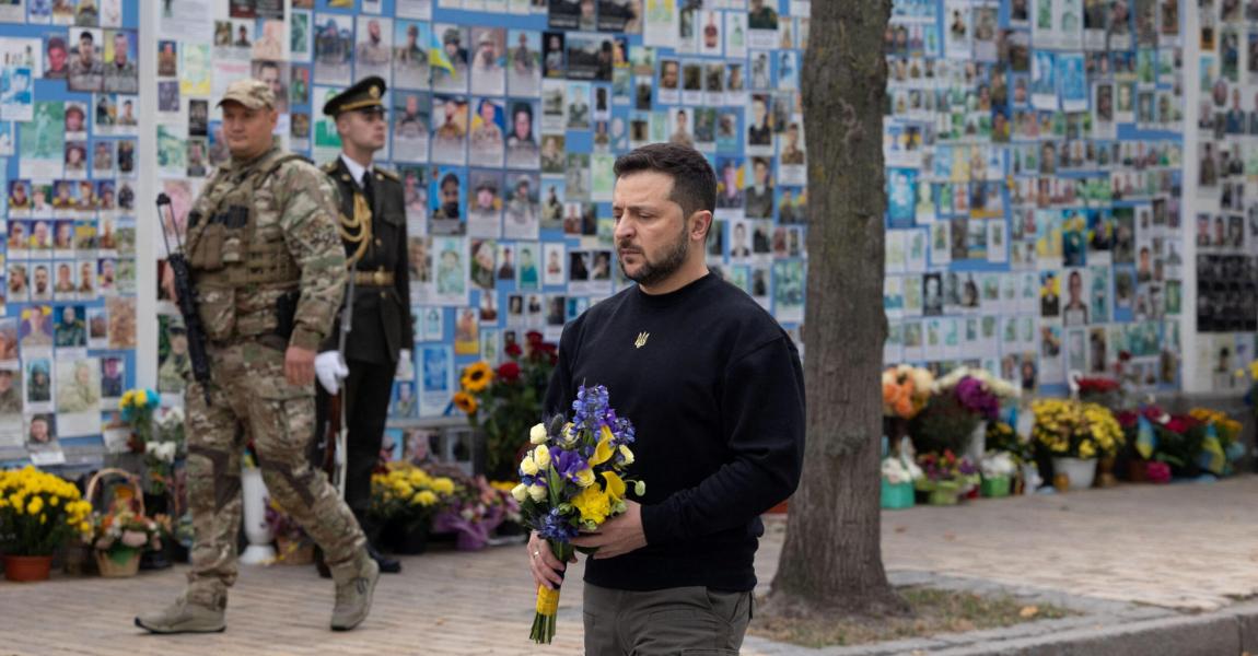 Ukrainian president volodymyr zelenskyy walking with a bouquet of flowers, guarded by soldiers, in front of a wall covered with photos of individuals.
