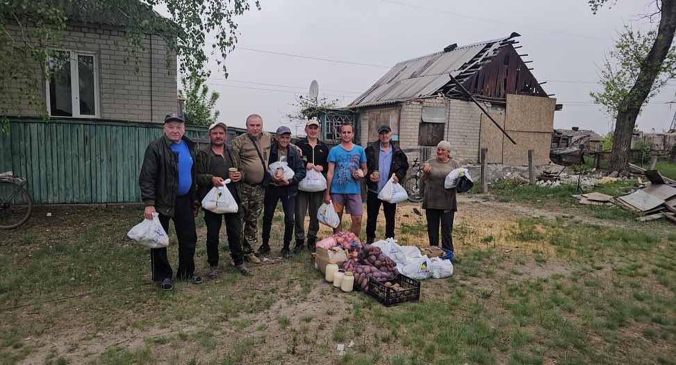 Group of adults standing outdoors with bags of groceries, surrounded by baskets of vegetables, with damaged buildings in the background, during a Hope for Ukraine Food Aid Distribution event.