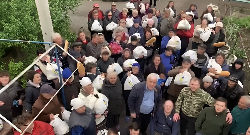 A group of people posing for a photo on an outdoor staircase during a Hope for Ukraine Food Aid Distribution event, most wearing white caps, with a diverse range of expressions.