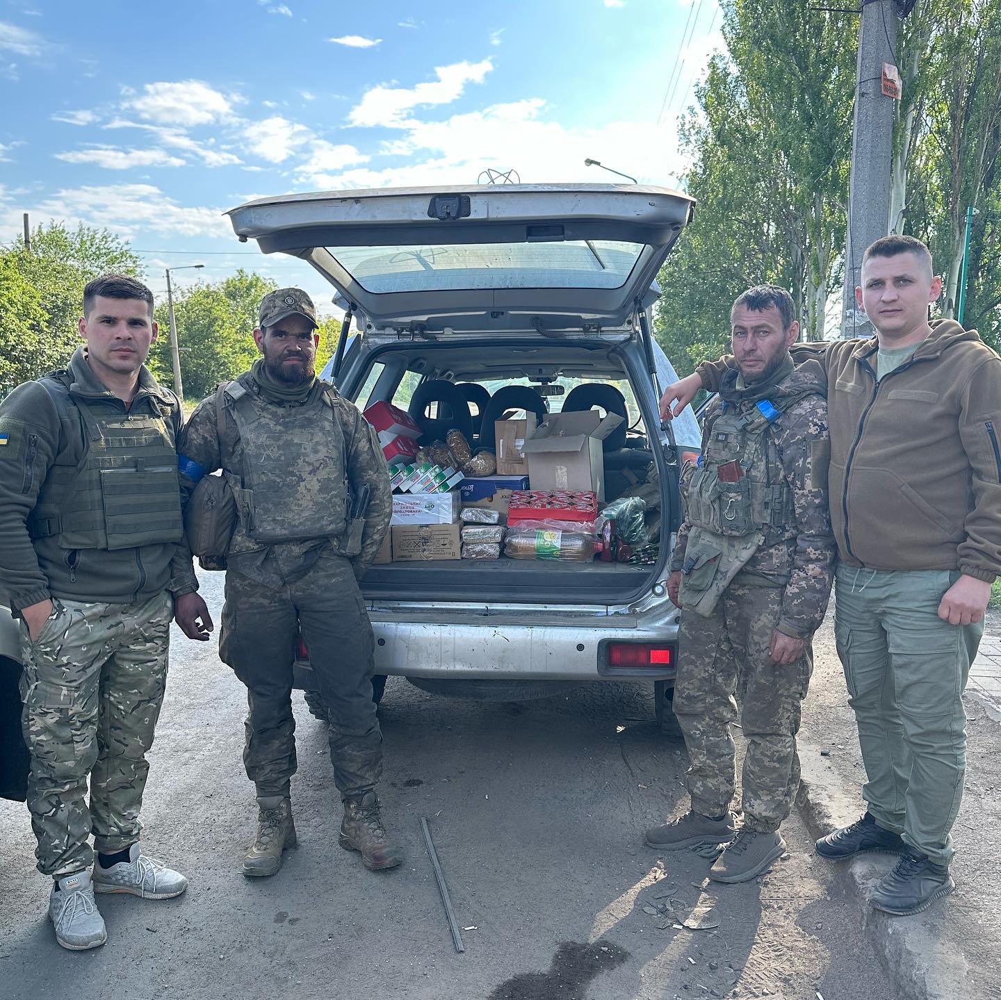 Four men in military attire stand outside by an open vehicle filled with boxes and supplies, offering Hope for Ukraine. Trees and a power pole are visible in the background.