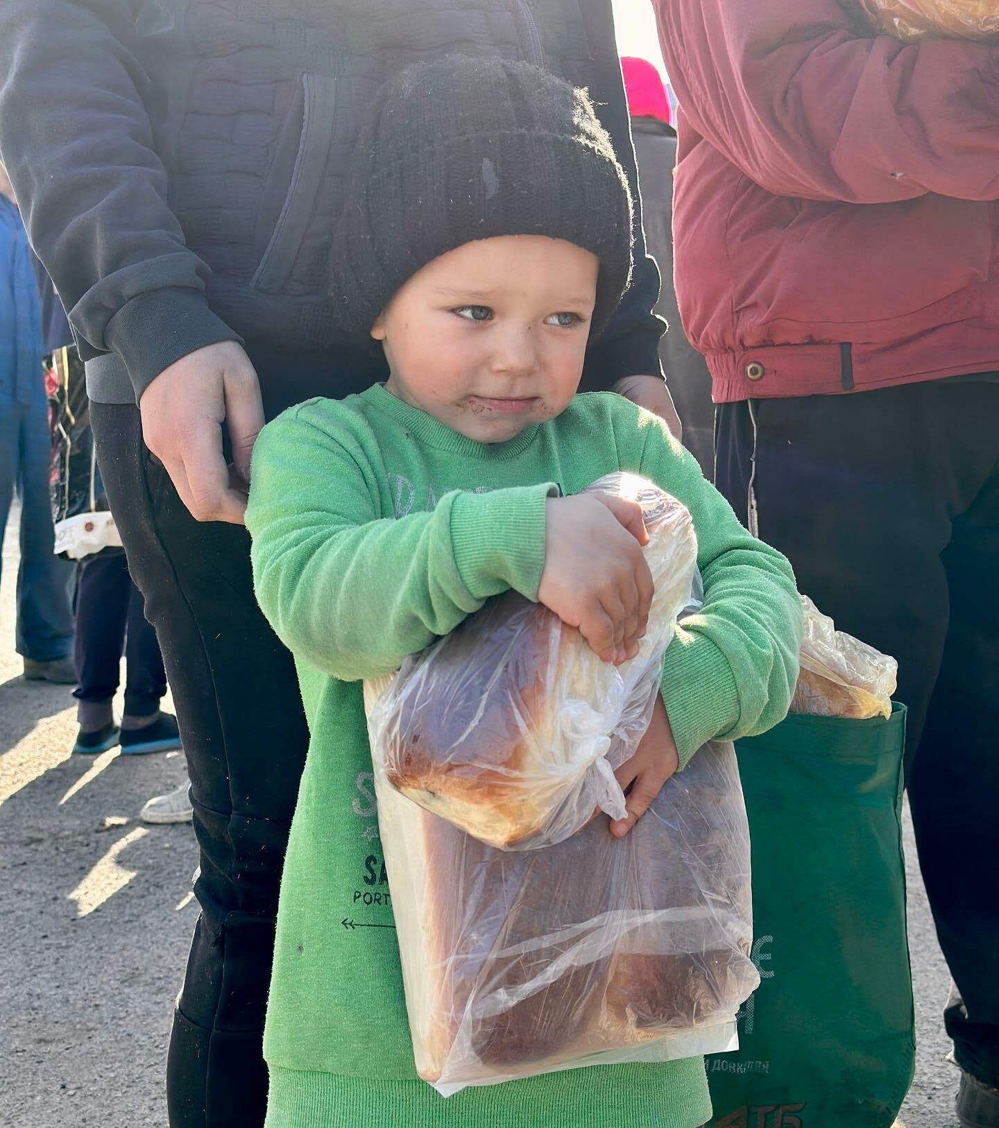 A young child in a green sweater and black beanie holds a large bag of bread while standing with an adult in a crowd.
