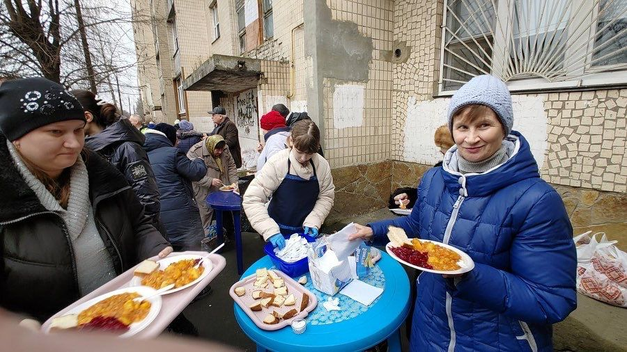 People standing outside in line receiving food on plates. A woman in a blue jacket and gray hat holds a plate and smiles at the camera. A person at a table serves food to others.