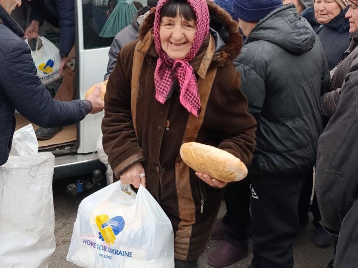 A woman in a headscarf and coat smiles while holding a loaf of bread and a plastic bag labeled "Hope for Ukraine" in a group of people near a van, symbolizing the impact when you donate to Ukraine charity.