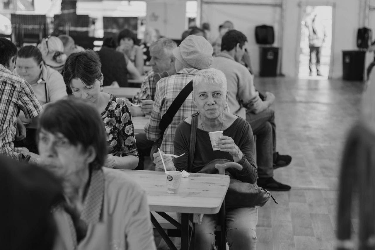 Inside a large tent, where food distribution brings hope for Ukraine, a group of people sit at tables. An older woman in the foreground holds a cup and spoon, looking directly at the camera.