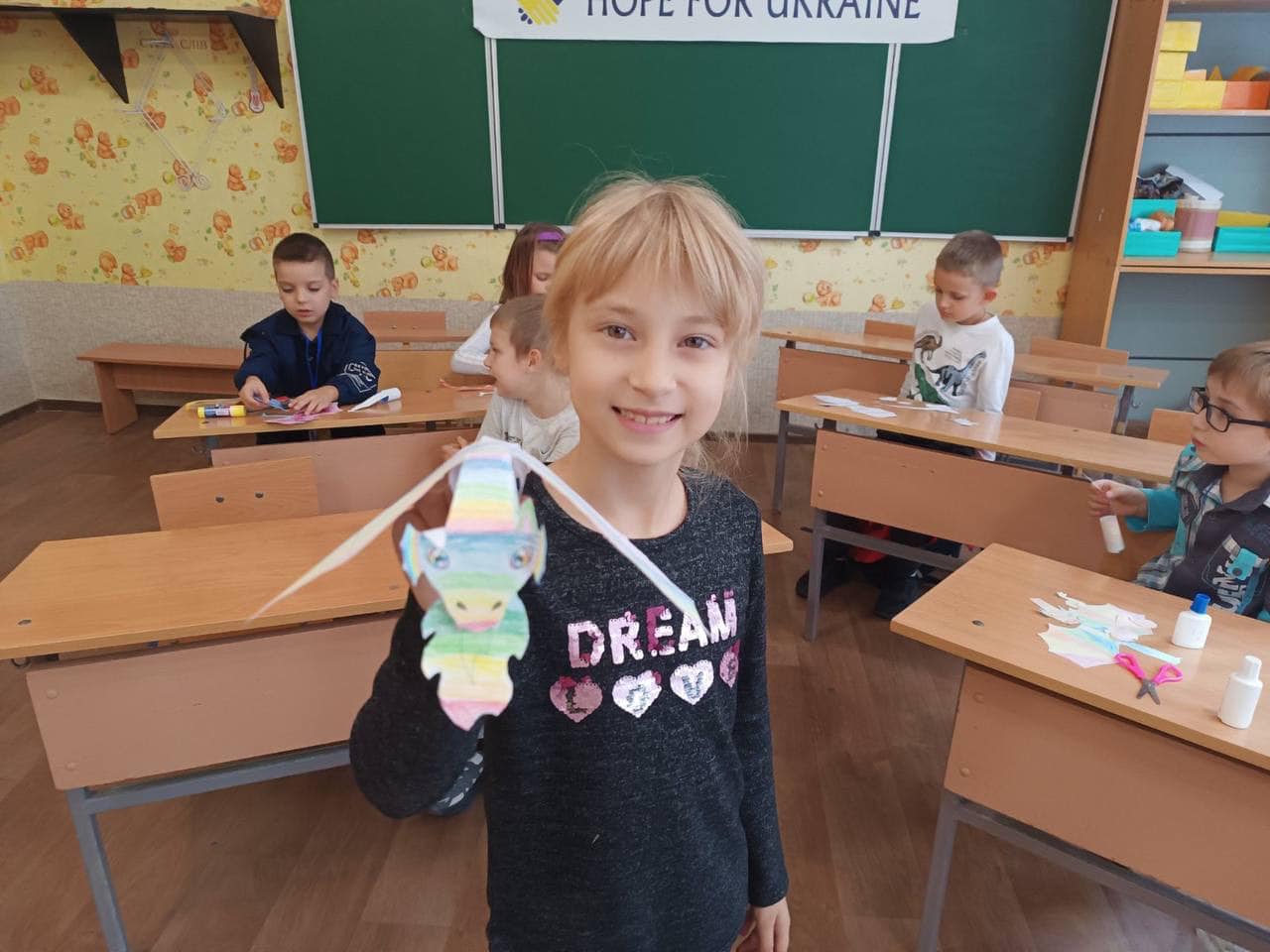 A young girl beams as she holds a colorful paper craft in a classroom, where the atmosphere is filled with hope. Other children, working at desks in the background, remind us that hope begins with a meal—a vital mantra guiding food distribution aid for Ukraine.