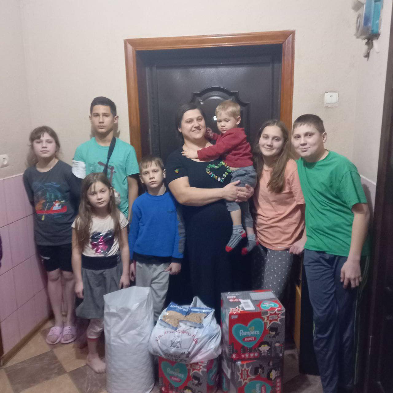 A group of people, including children and an adult holding a toddler, stand indoors, surrounded by bags and boxes filled with resources from the children's educational support program.