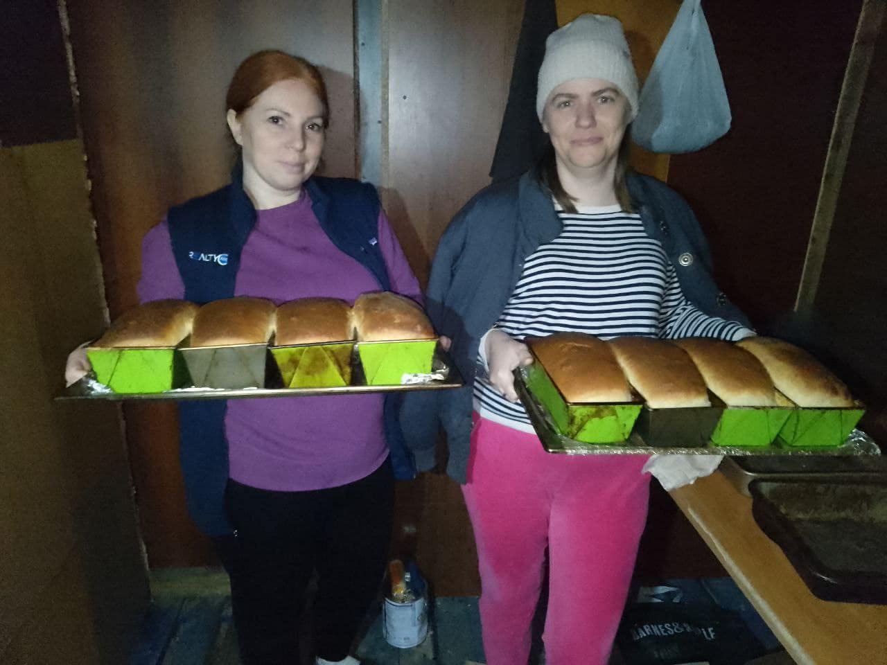 Two people holding trays of freshly baked bread in a dimly lit room, with the warmth reflecting as if painting smiles on glass.