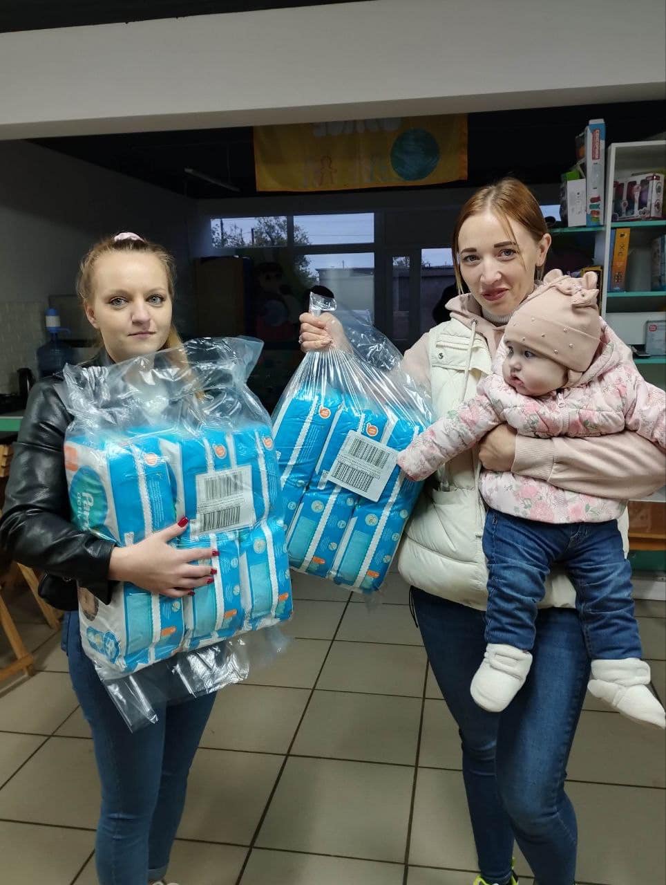 Two women, embodying the spirit of Delivering Hope to Orphans, clutch packs of diapers, one gently balancing a toddler on her hip. They stand indoors, surrounded by shelves filled with essentials ready to bring comfort and care.