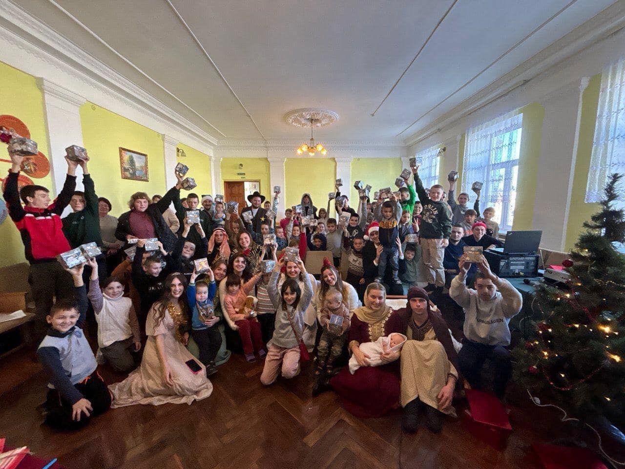 A large and joyous group gathers indoors, posing for a photo amidst festive decorations. Some hold up objects symbolizing the theme 