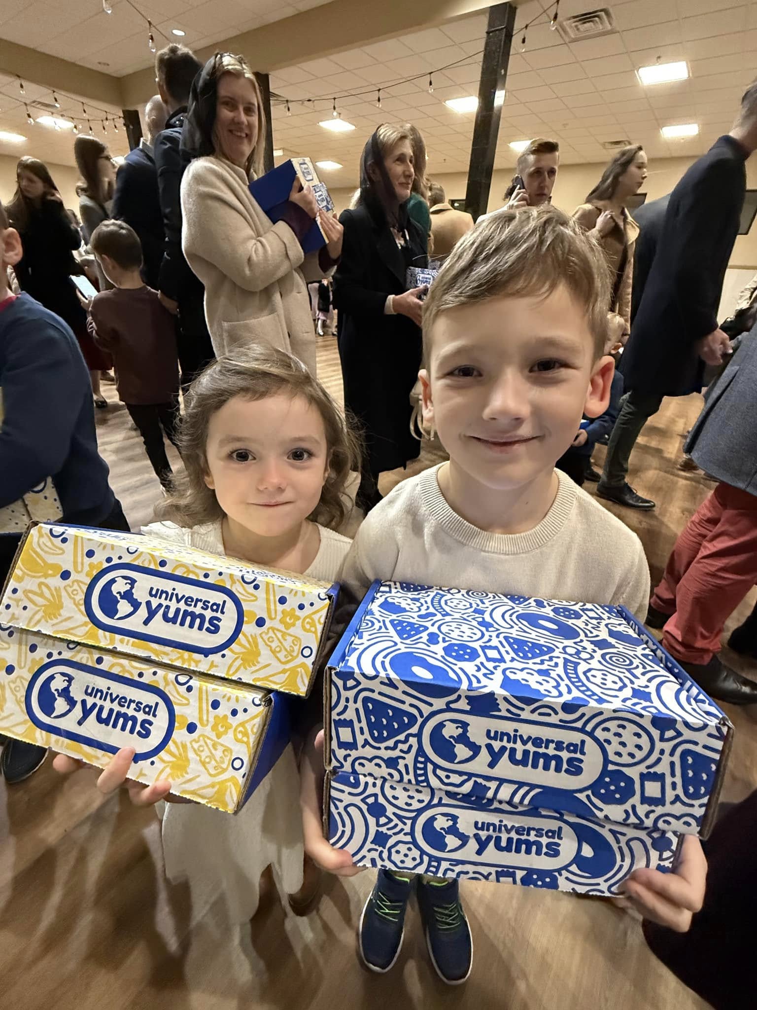 Two children, filled with the joy of Christmas, hold Universal Yums boxes at an indoor event bustling with people in the background.