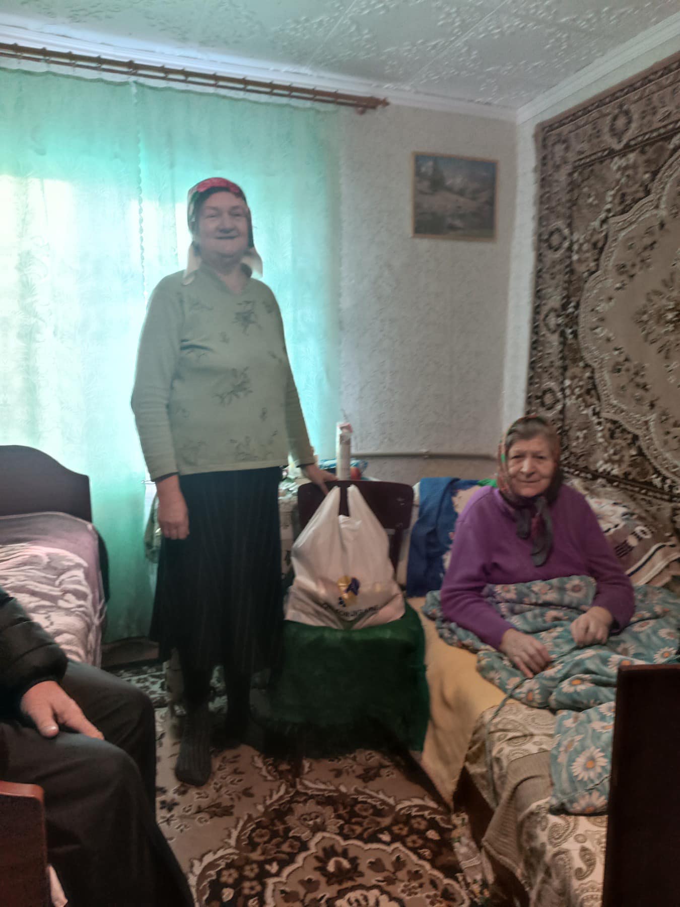 In a warmly decorated room with patterned carpet and walls, two elderly women share a moment. One stands beside a table with a bag, while the other sits on the bed, reflecting on their recent gathering at Meals of Hope in Lviv.