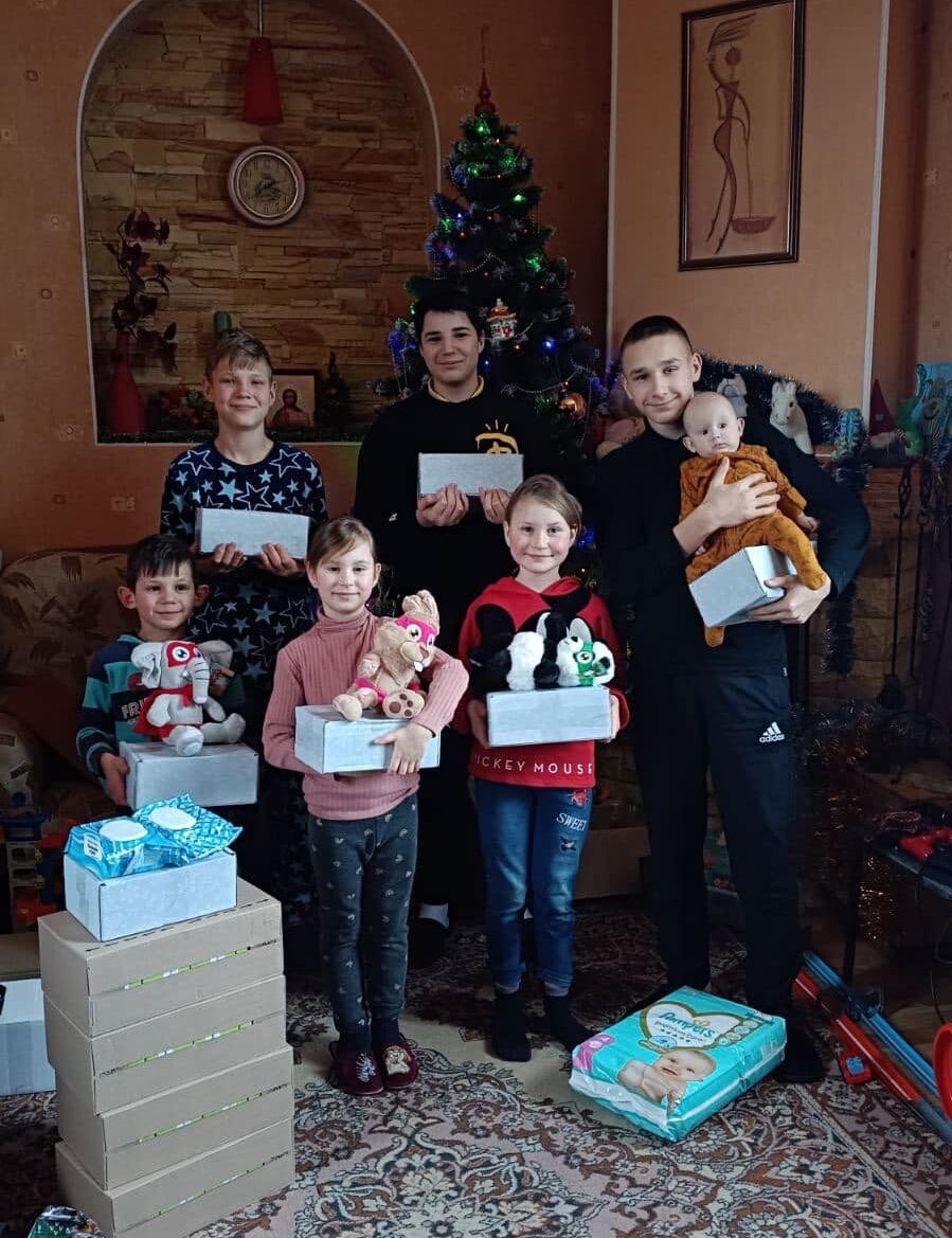 A group of six people, four children and two adults, joyfully stand in a living room brimming with gifts. A beautifully decorated Christmas tree gleams in the background, perfectly capturing the spirit of spreading holiday joy.