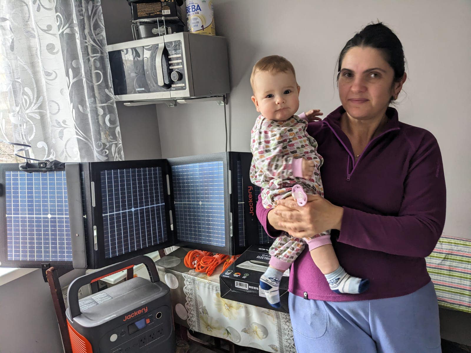 In a room illuminated with hope, a woman cradles her baby beside a portable solar panel setup, where a power station hums softly among the electronics on the table. It's truly light in the darkness.