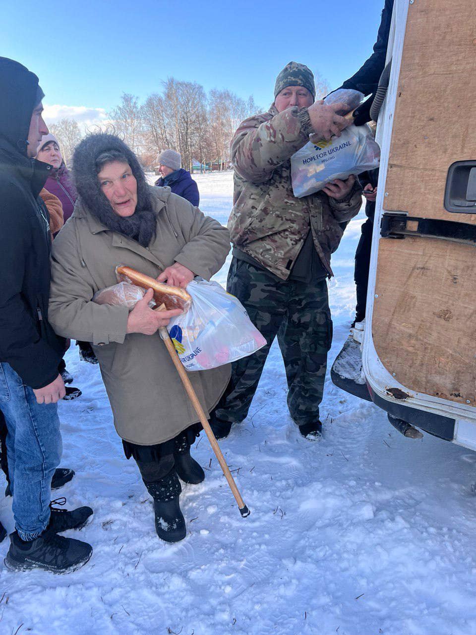 People receive supplies from a vehicle in a snowy outdoor setting. An older woman with a cane holds bags of goods, while a man in camouflage hands down food kits for Kharkiv families.