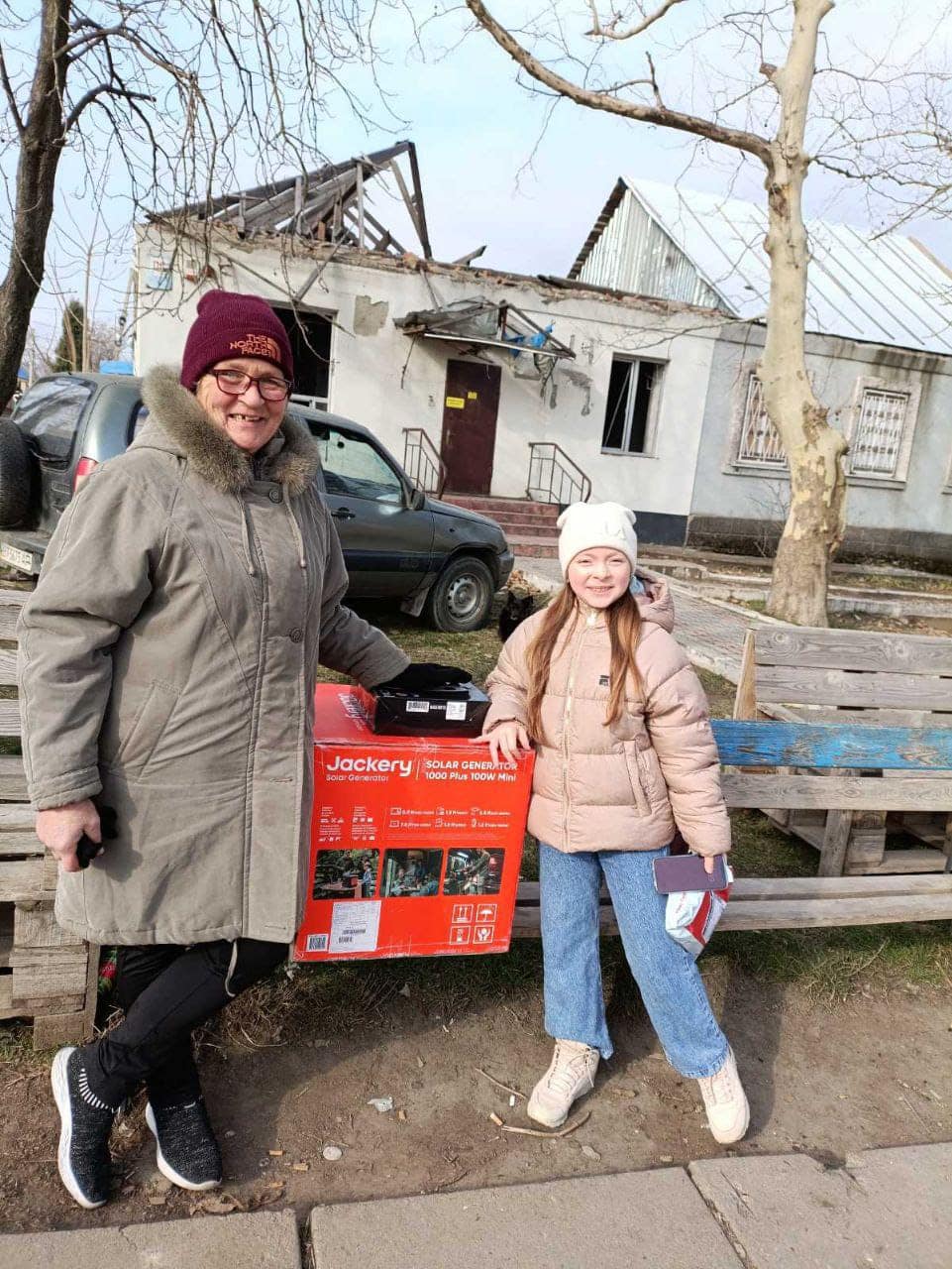 Two people stand outdoors in front of a damaged building, a beacon of hope amidst the rubble. One holds a Jackery solar generator box—a true light in the darkness. They are both dressed warmly and smiling, embracing resilience even in challenging times.