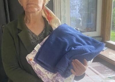 An older woman wearing glasses and a headscarf stands indoors, holding folded blankets in her arms next to a bag of groceries on a table—a scene reflecting the impact of Nourishing Northern Ukraine.