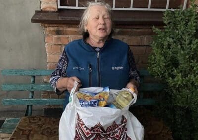 An elderly woman sits on a bench outside, holding a shopping bag with food items in her lap and smiling with her eyes closed, reflecting the warmth of Nourishing Northern Ukraine.