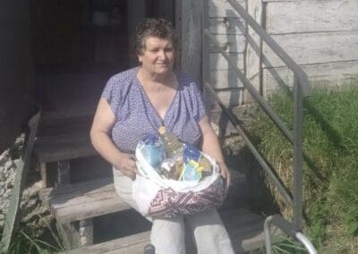 A woman sits on wooden steps outside a weathered building in Northern Ukraine, holding a bag of nourishing groceries and using a walker nearby.