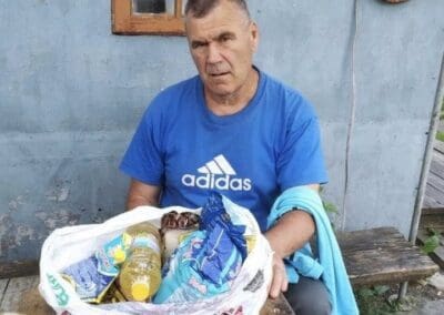 A man in a blue Adidas shirt sits at a wooden table outdoors, sharing a moment of Nourishing Northern Ukraine with a bag of groceries including oil and packaged food in front of him.