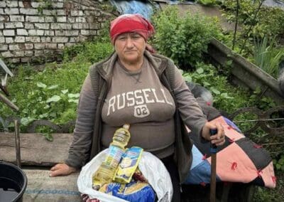 An elderly woman with a red headscarf stands outdoors, holding a cane beside a bag of groceries on a table—an image reflecting the spirit of Nourishing Northern Ukraine amid greenery and a brick wall.