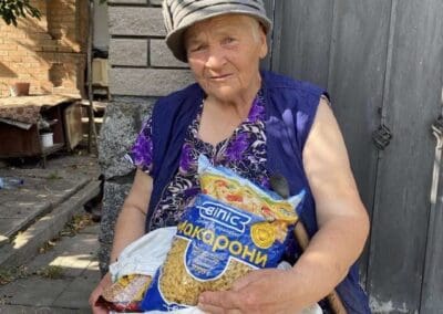 An elderly woman in a hat and purple vest sits outside holding a cane, a bag of pasta, and a white bag with a patterned cloth—reflecting the spirit of Nourishing Northern Ukraine.