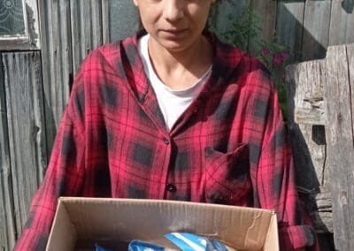 A woman in a red plaid shirt stands outside holding a cardboard box filled with multiple packages of pasta, sharing Kindness Across Kriviy Rih.