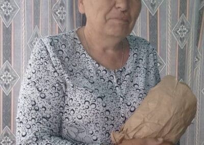 An older woman with short gray hair, wearing a patterned shirt, stands indoors holding a brown paper-wrapped package in front of a geometric-patterned curtain—one of many Sumy Families Nourished through thoughtful support.