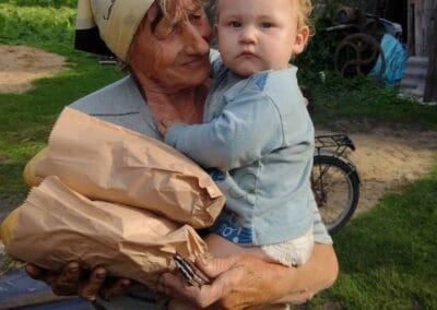 An elderly woman holds a baby and two loaves of bread wrapped in paper, standing outside near an old wooden building—illustrating how Sumy Families Nourished brings comfort and sustenance across generations.