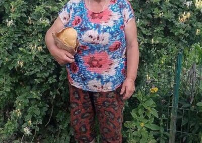 A woman in patterned clothing stands outdoors in front of a hedge, holding a large mushroom and smiling at the camera—reflecting how Sumy Families Nourished celebrates nature’s bounty.