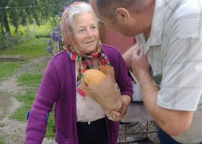An elderly woman with a cane holds a loaf of bread wrapped in paper while standing outside, as a man speaks to her—one of the many moments where Sumy Families Nourished brings hope to the community.