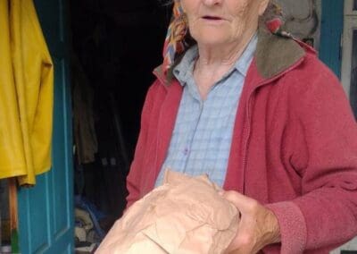 An older woman in a headscarf and red jacket stands near a doorway, holding a crumpled brown paper bag and a box of mushrooms—one of many Sumy Families Nourished through community support.