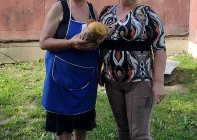 Two women stand outside on grass; the older woman on the left, part of Sumy Families Nourished, wears a blue apron and holds a loaf of bread while the younger woman stands beside her, both smiling at the camera.