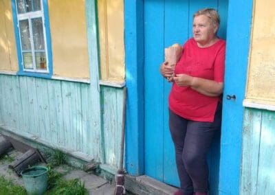 A woman in a red shirt stands in a blue doorway, holding a wooden object. Sumy Families Nourished, she tends to her rustic wooden house, with a broom and green bucket close by.