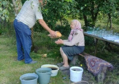 An elderly woman sits on a bench outdoors as a man hands her a melon, helping keep Sumy families nourished. Several buckets, jars, and a basket rest on the grass nearby, with trees and greenery in the background.