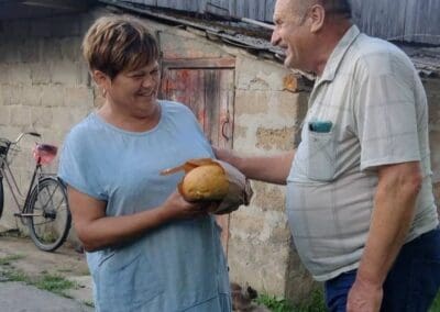 A woman in a light blue dress holds a round loaf of bread, smiling at a man in a plaid shirt outside a rustic building with a bicycle—a scene reflecting how Sumy Families Nourished celebrate simple joys together.
