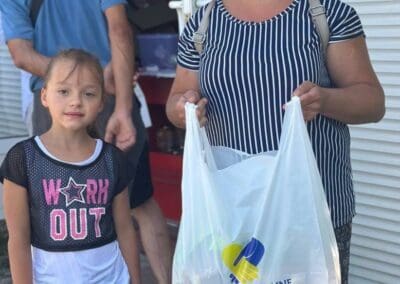 A woman holds a "Hope for Ukraine" bag, symbolizing Food Aid for Northern Ukraine, as she stands next to a young girl; other people are visible in the background near a building.