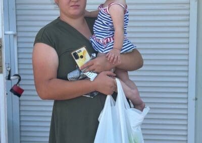A woman in a green dress holds a young child with a pacifier, a smartphone, keys, and a plastic shopping bag—perhaps containing Food Aid for Northern Ukraine—while standing outside near a white shuttered door.