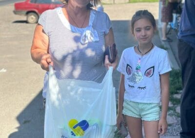 A woman and a girl stand outdoors holding a plastic bag with the "Home For Ukraine" logo, part of Food Aid for Northern Ukraine. Buildings, a red car, and other people are visible in the background.