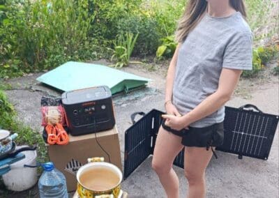 A woman stands outdoors beside a portable power station, solar panel, water jug, and pot on a small stove—Powering Hope Near Frontlines amid the greenery and pavement in the background.