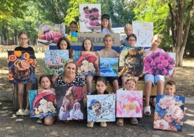 A group of children and one adult sit outdoors holding paintings of flowers, animals, and desserts, with a "Hope for Ukraine" sign in the background, enjoying Creative Safe Spaces to express themselves.