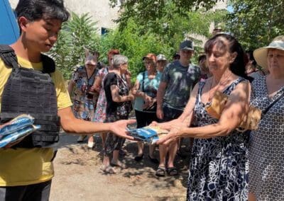 A man in a protective vest from Helping Hands in Kharkiv distributes supplies to a group of people standing outdoors in the shade, with trees and a damaged building in the background.