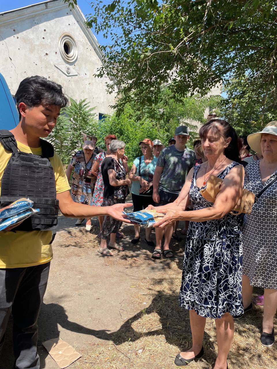 A man in a protective vest from Helping Hands in Kharkiv distributes supplies to a group of people standing outdoors in the shade, with trees and a damaged building in the background.