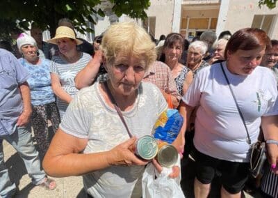 An elderly woman stands in a crowd outdoors, holding canned food and packaged goods from Helping Hands in Kharkiv in one hand and a plastic bag in the other.