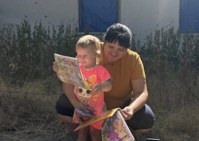A woman kneels beside a young girl standing on a toy wheelbarrow, both examining colorful books outdoors near a building with boarded windows—a touching moment captured by Helping Hands in Kharkiv.