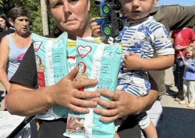 A woman holds a young boy, two packs of diapers, and a toy truck—standing among a group of people outdoors on a sunny day, where Firewood for Survival is also being distributed.