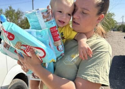 A woman carrying a young child and several packs of diapers stands outside near a white van on a sunny day, ready with essentials like firewood for survival.